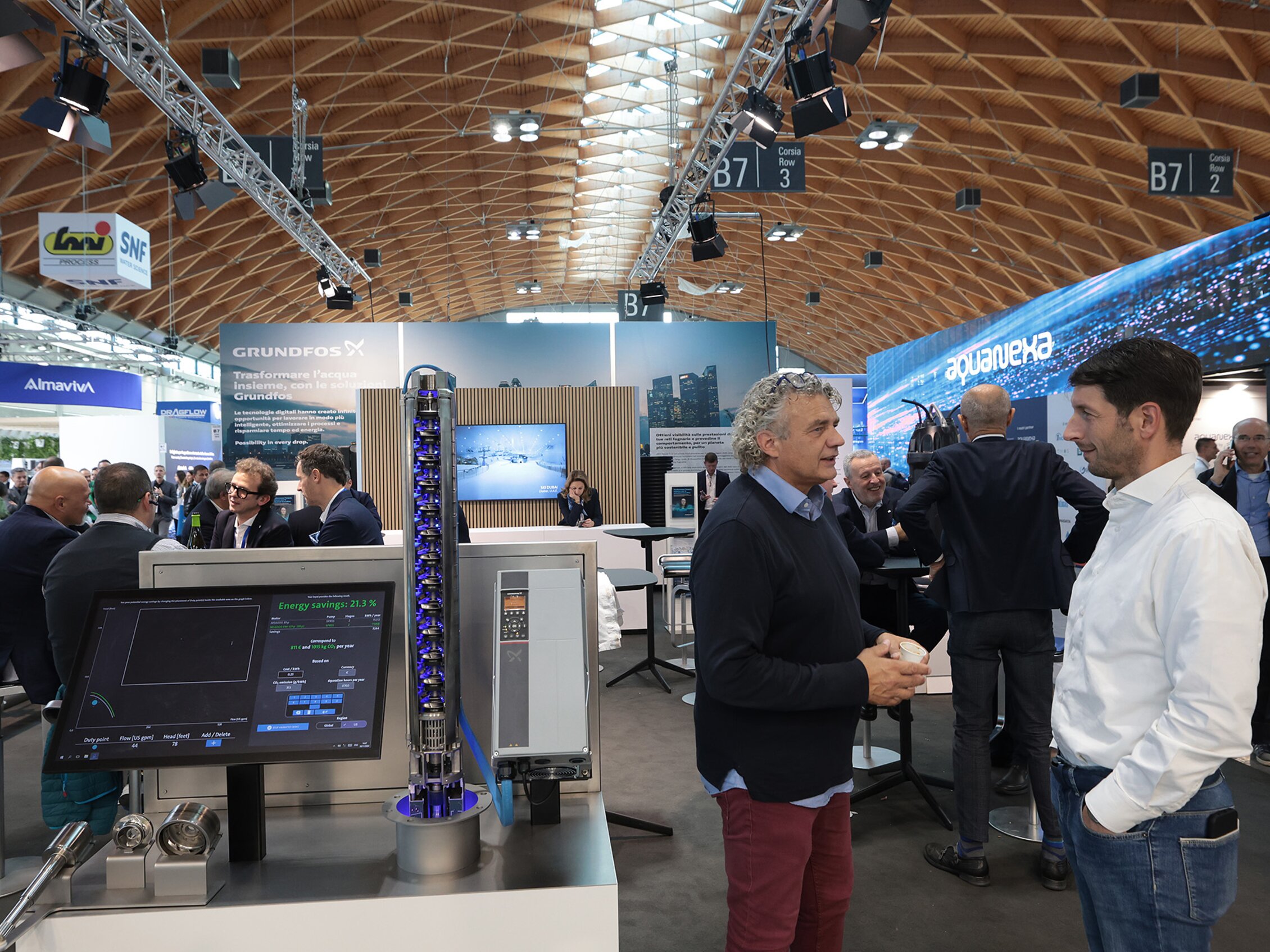 partecipanti a Ecomondo parlano fra di loro circondati dai macchinari e dai display degli stand di Almaviva, Grundfos e aquaVEXA